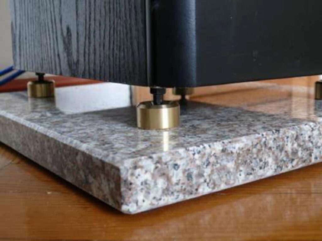 Base de piedra natural pulida colocada bajo un altavoz, con patas metálicas de desacoplamiento apoyadas sobre la superficie de granito.